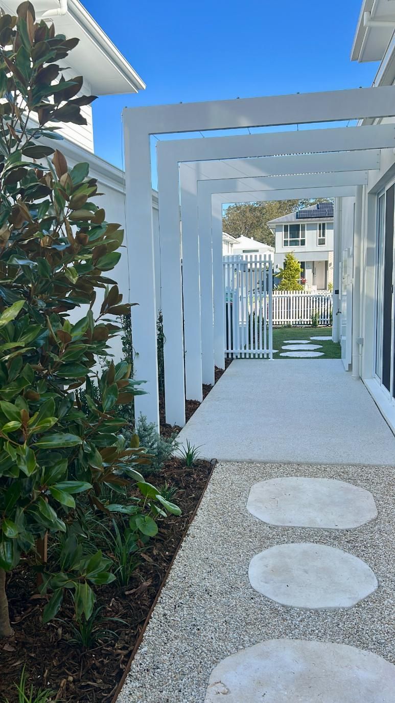 A white pergola leads along a pebble path with round stepping stones toward a white fenced yard on a sunny day.