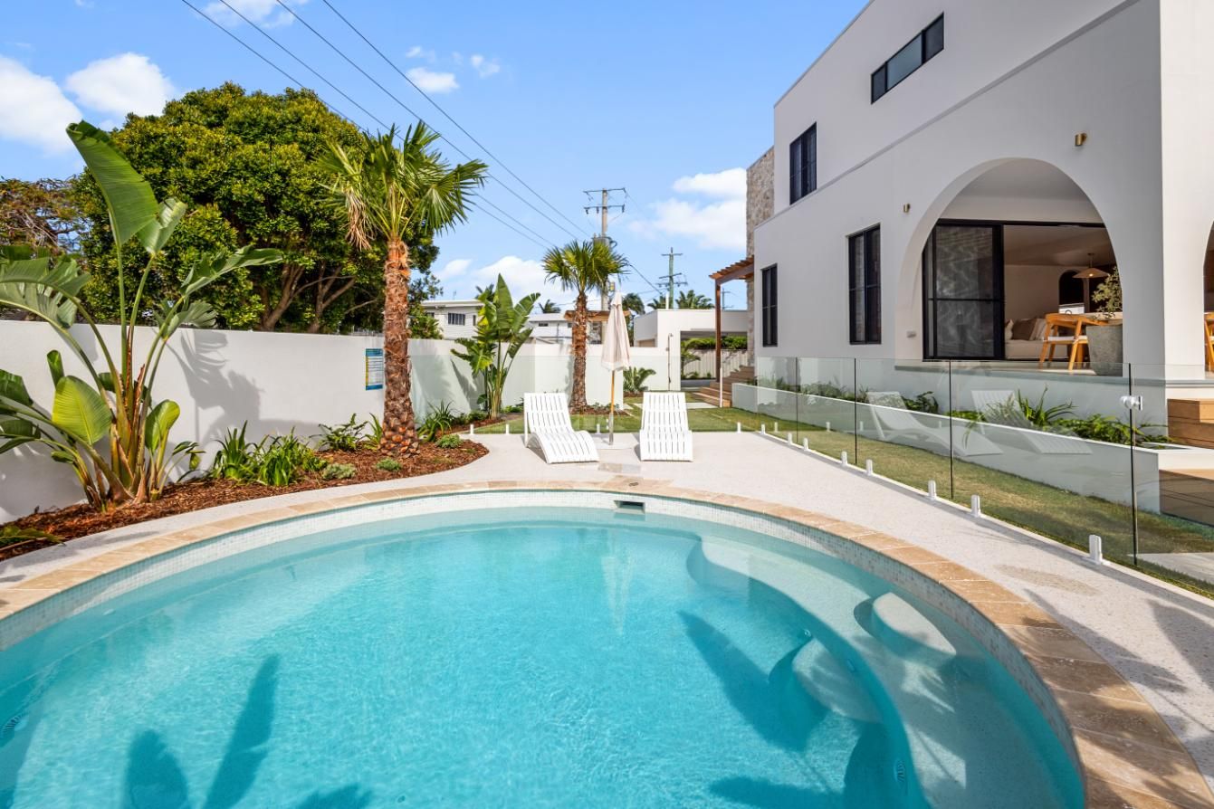 A bright backyard with a clear swimming pool, two white lounge chairs, palm trees, and a modern white house with an arch.