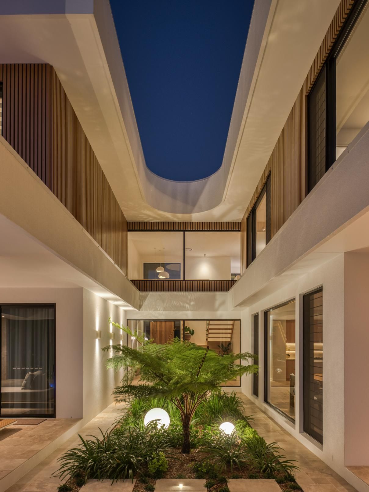A modern courtyard with a central garden at night, surrounded by illuminated two-story walls with wood and glass accents.