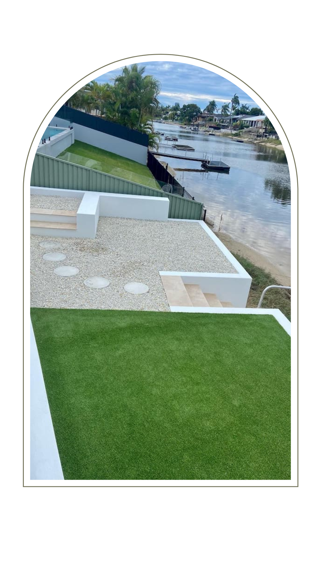 A tiered backyard with green grass, a gravel patio with stepping stones, and white retaining walls overlooking a river.