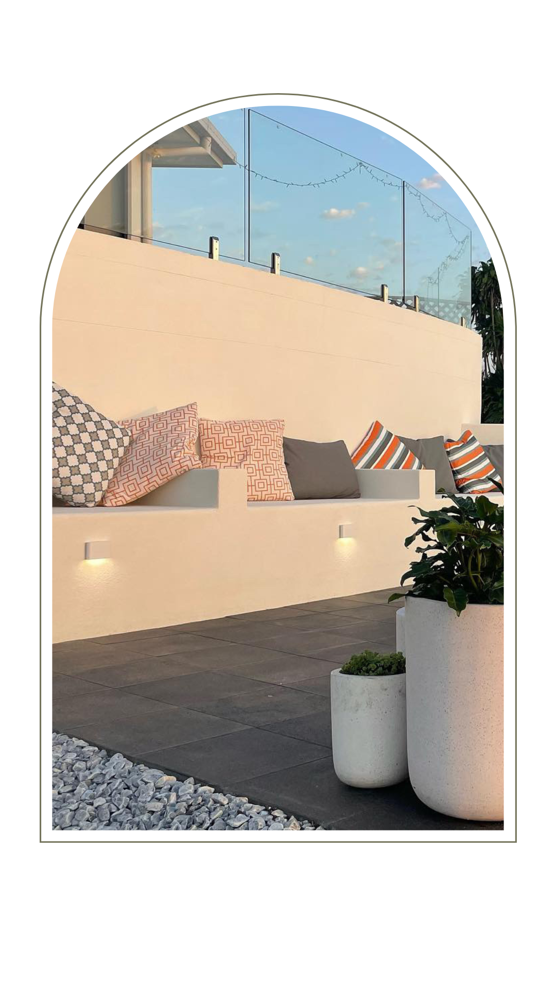An outdoor patio with a beige seating wall lined with decorative pillows, lit by wall-mounted lights and potted plants.