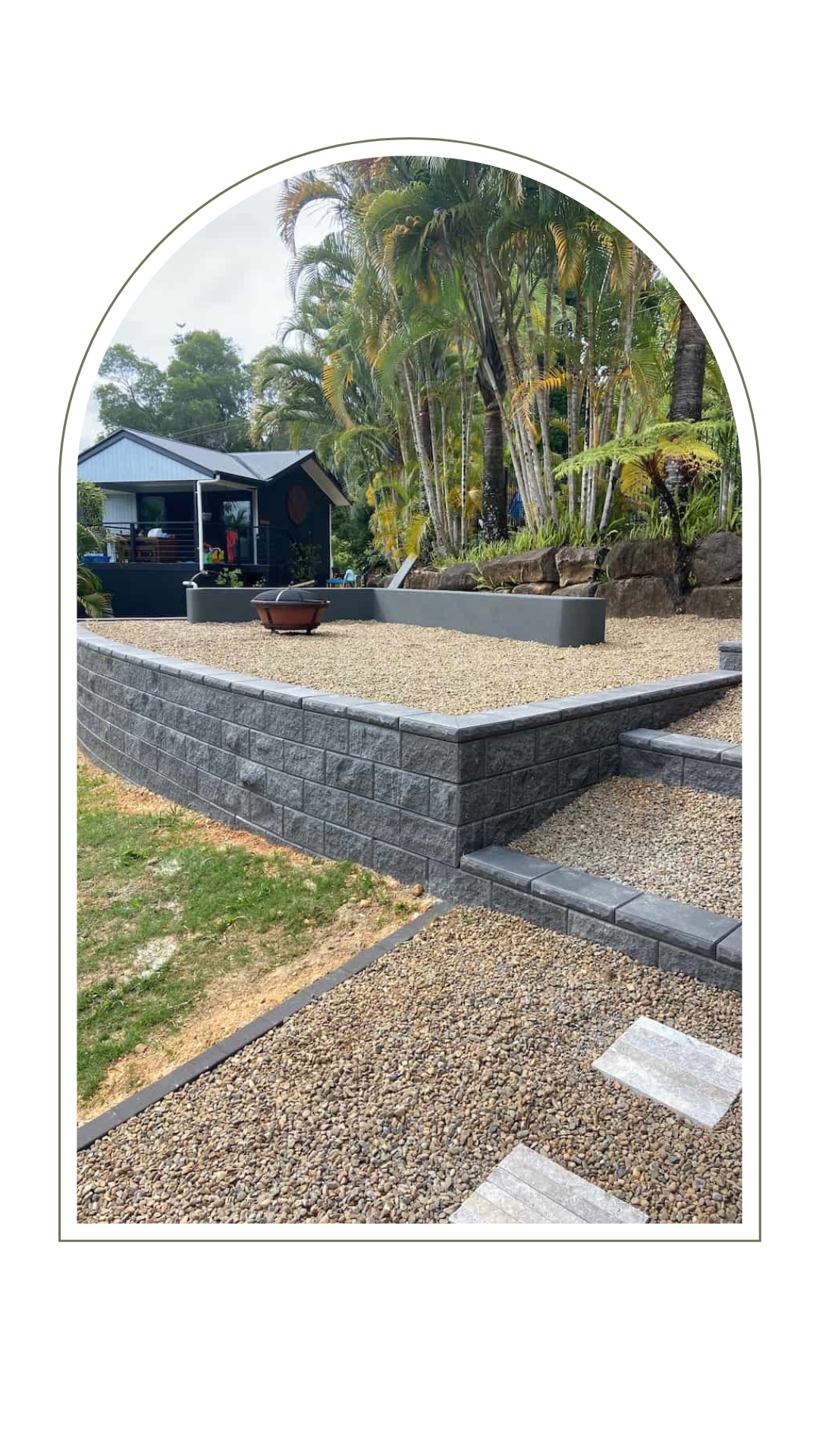 A tiered outdoor patio area with dark gray stone retaining walls, gravel landscaping, and a fire pit near a small cabin.