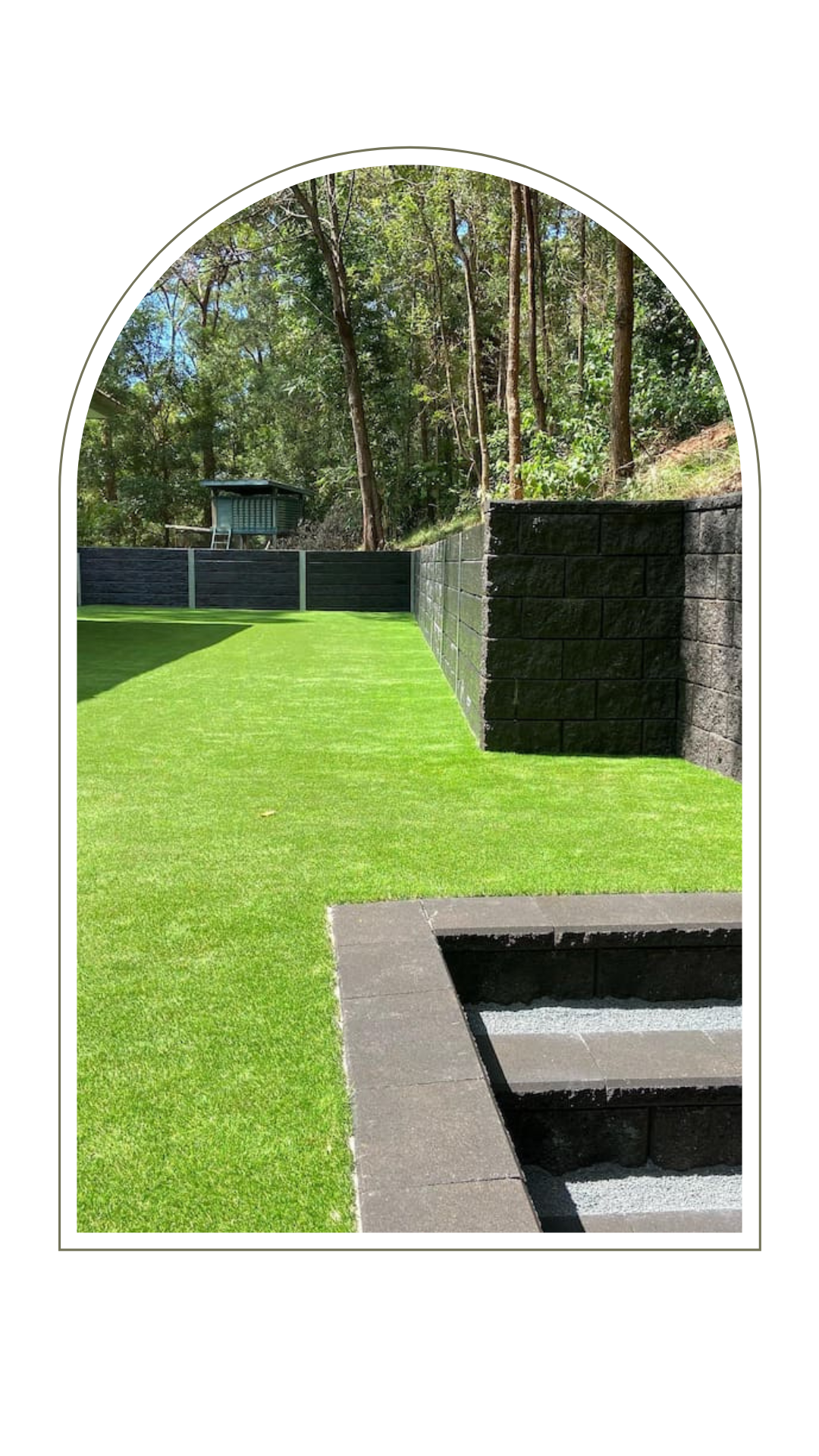 A manicured lawn bordered by a tall stone retaining wall and a staircase, with trees in the background.