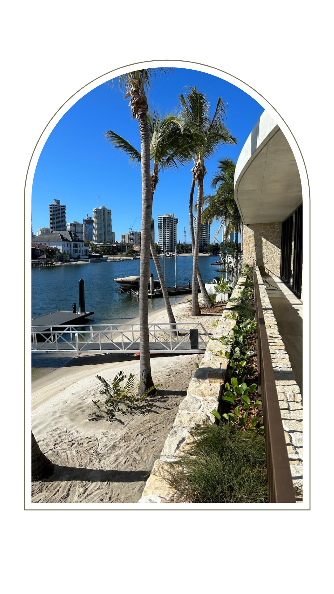 A waterfront view of a canal with palm trees, a sandy bank, and city skyline buildings under a clear blue sky.