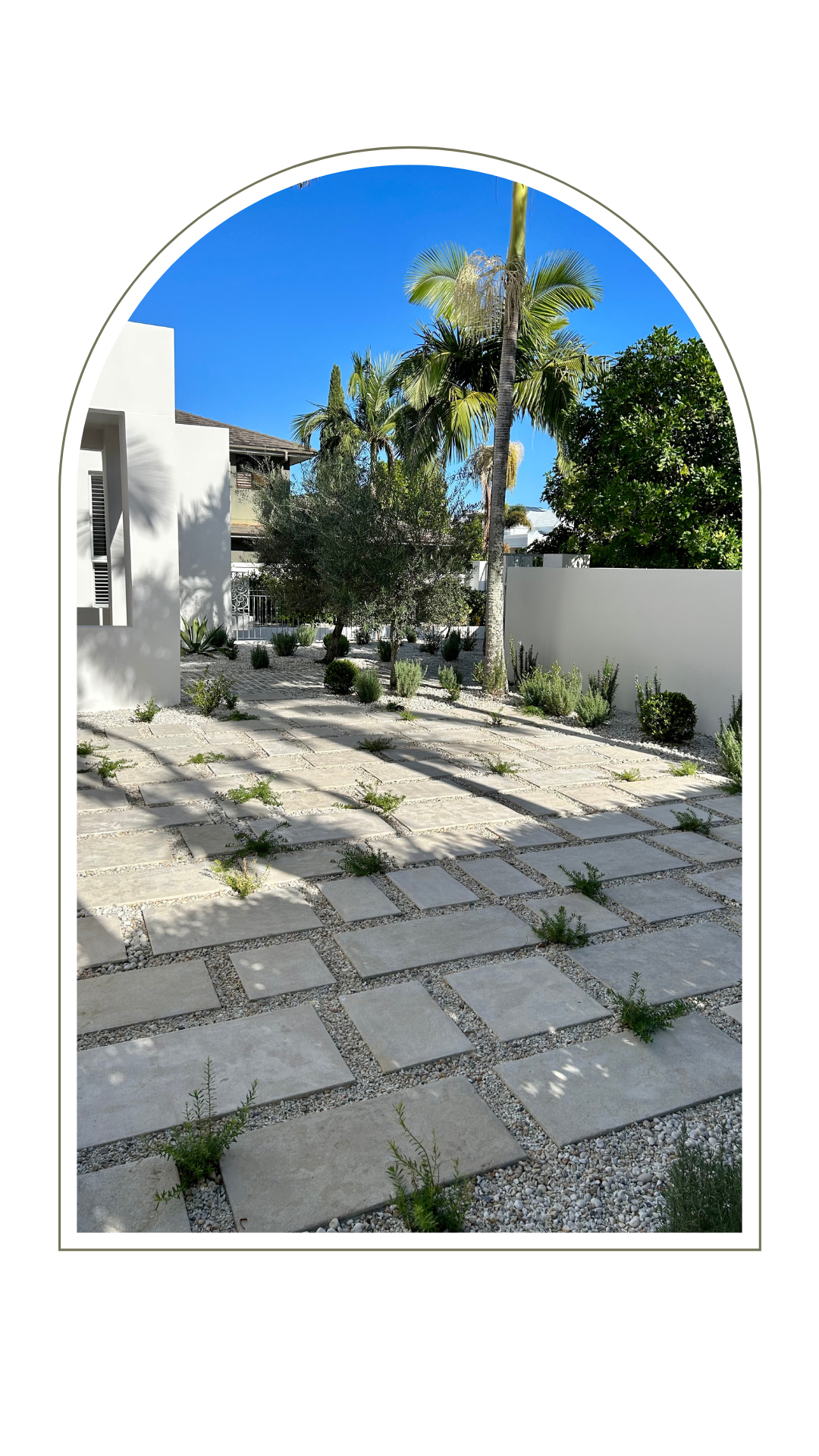 A minimalist outdoor patio with grey rectangular pavers surrounded by white gravel, palm trees, and a white wall.