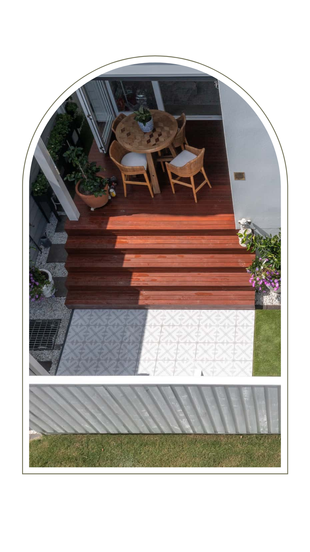 A high-angle view of a wooden deck with a table and chairs, leading down steps to a patio and a grass lawn.