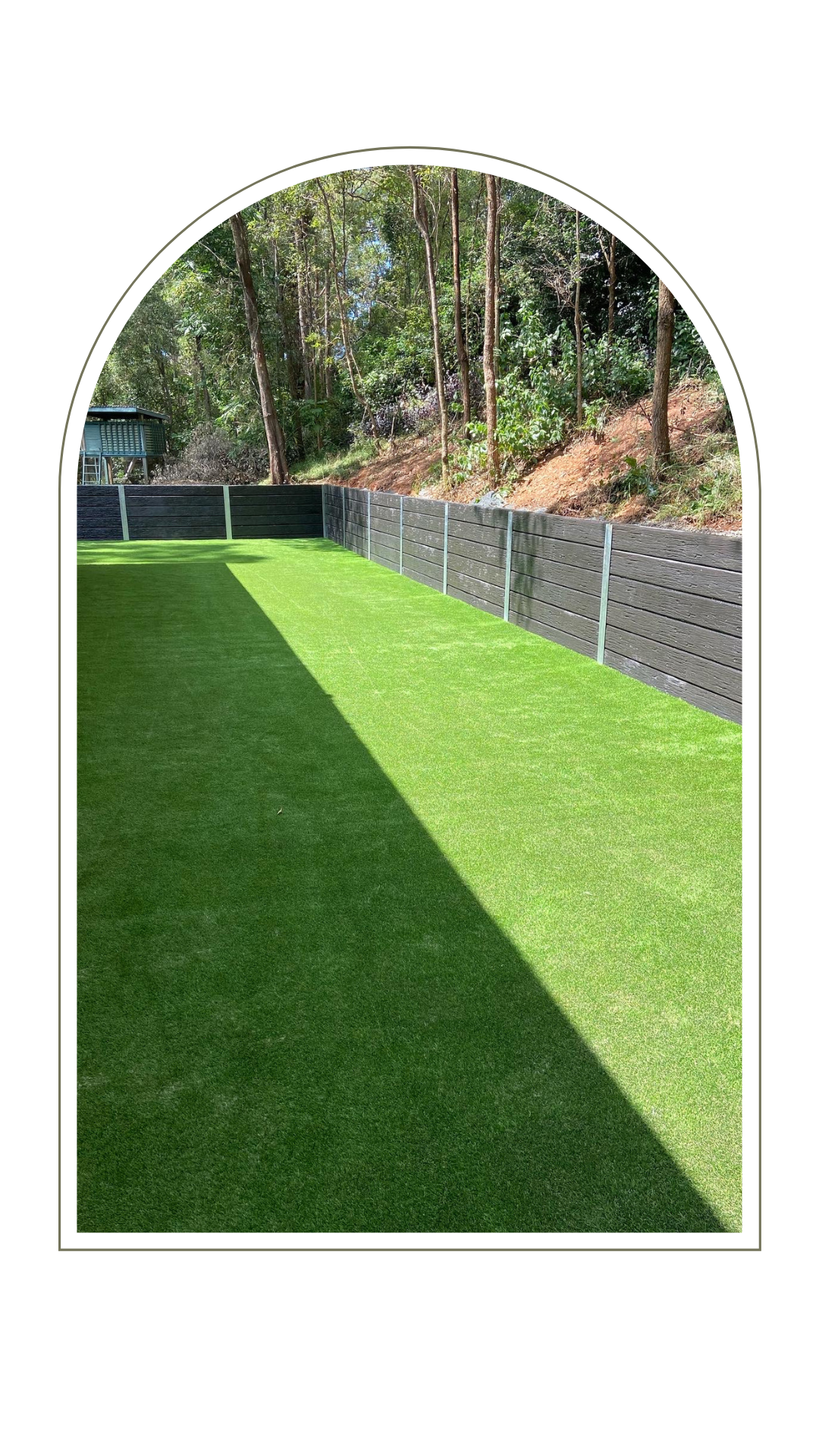 A backyard lawn split by a diagonal shadow, bordered by a black slatted fence and trees, set in an arched frame.