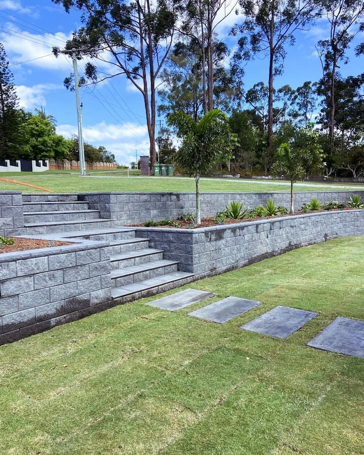 A stone retaining wall with integrated steps leading to an upper lawn, set in a sunny backyard with trees and grass.