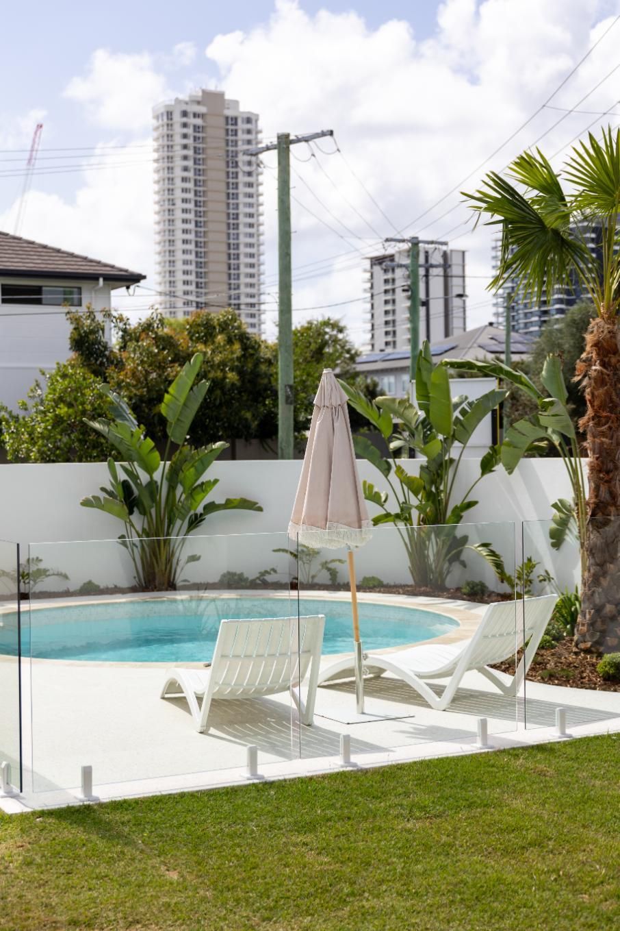 Two white lounge chairs and a closed parasol by a small circular pool in a backyard with city high-rises in the distance.
