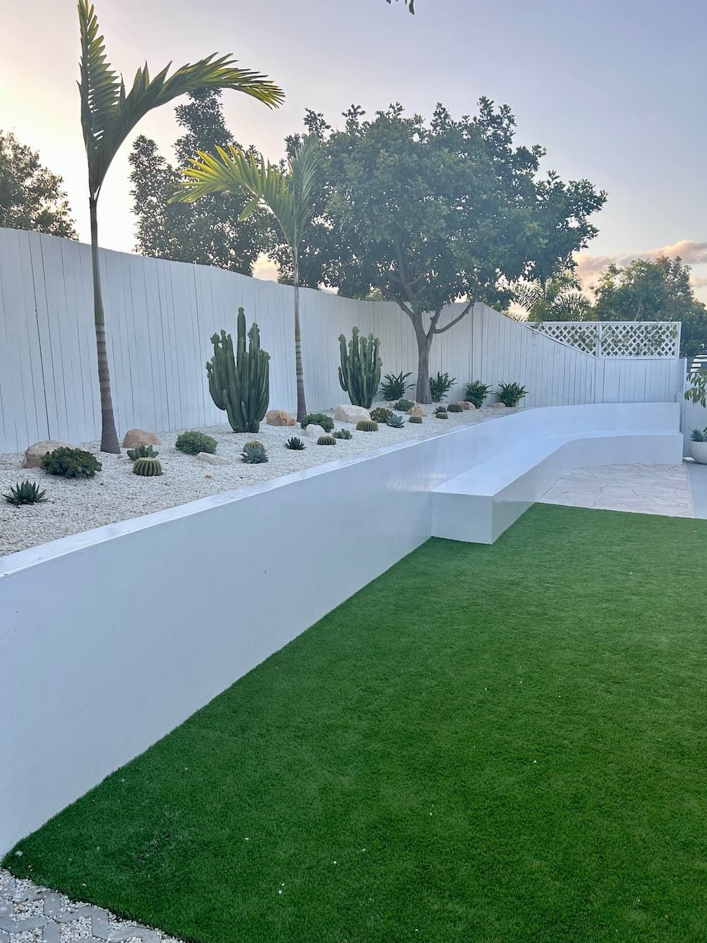 a white fence surrounds a garden with cactus and palm trees and artificial turf