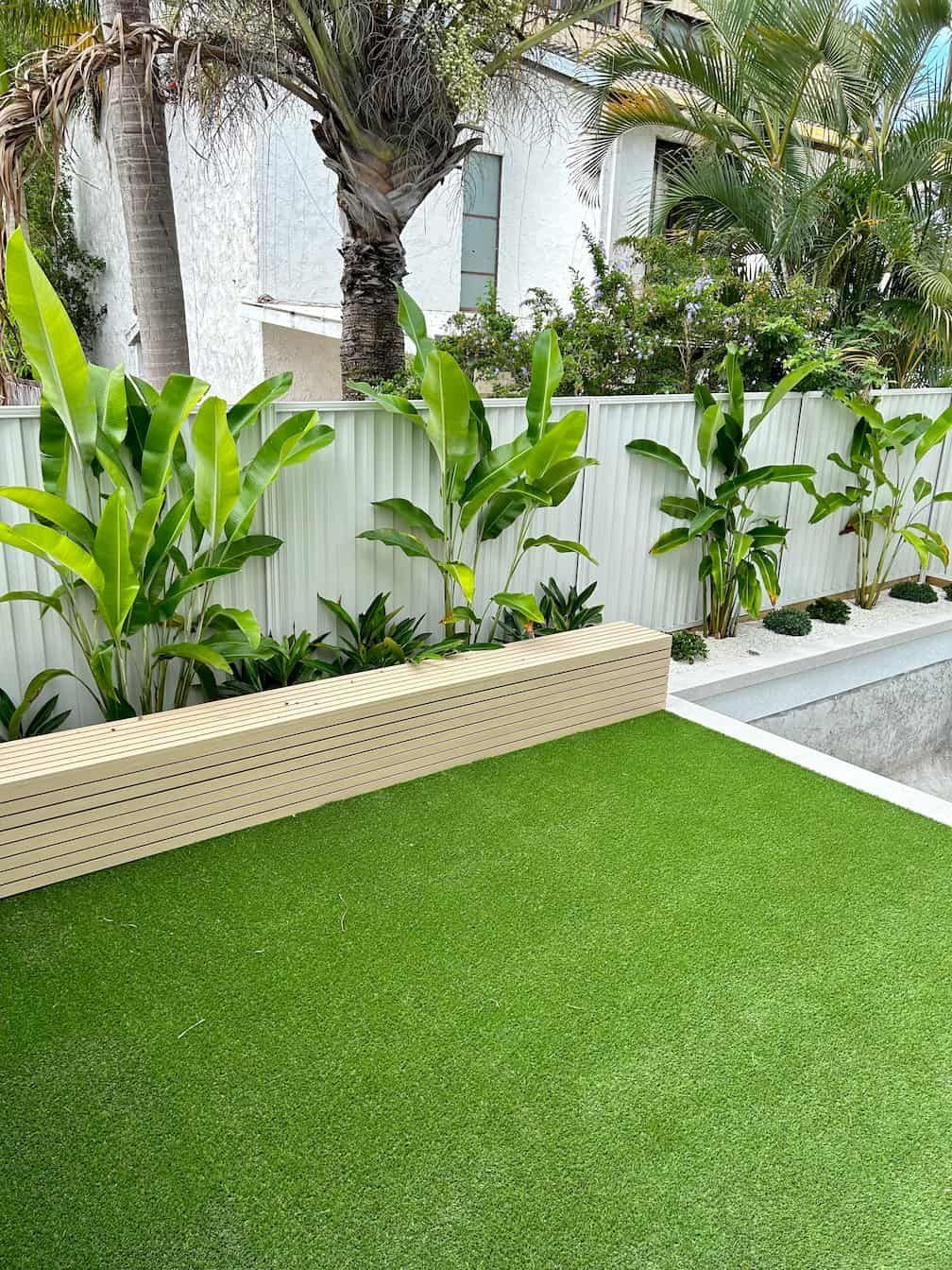 A bright green artificial lawn sits before a beige retaining wall and white fence, featuring lush green tropical plants.
