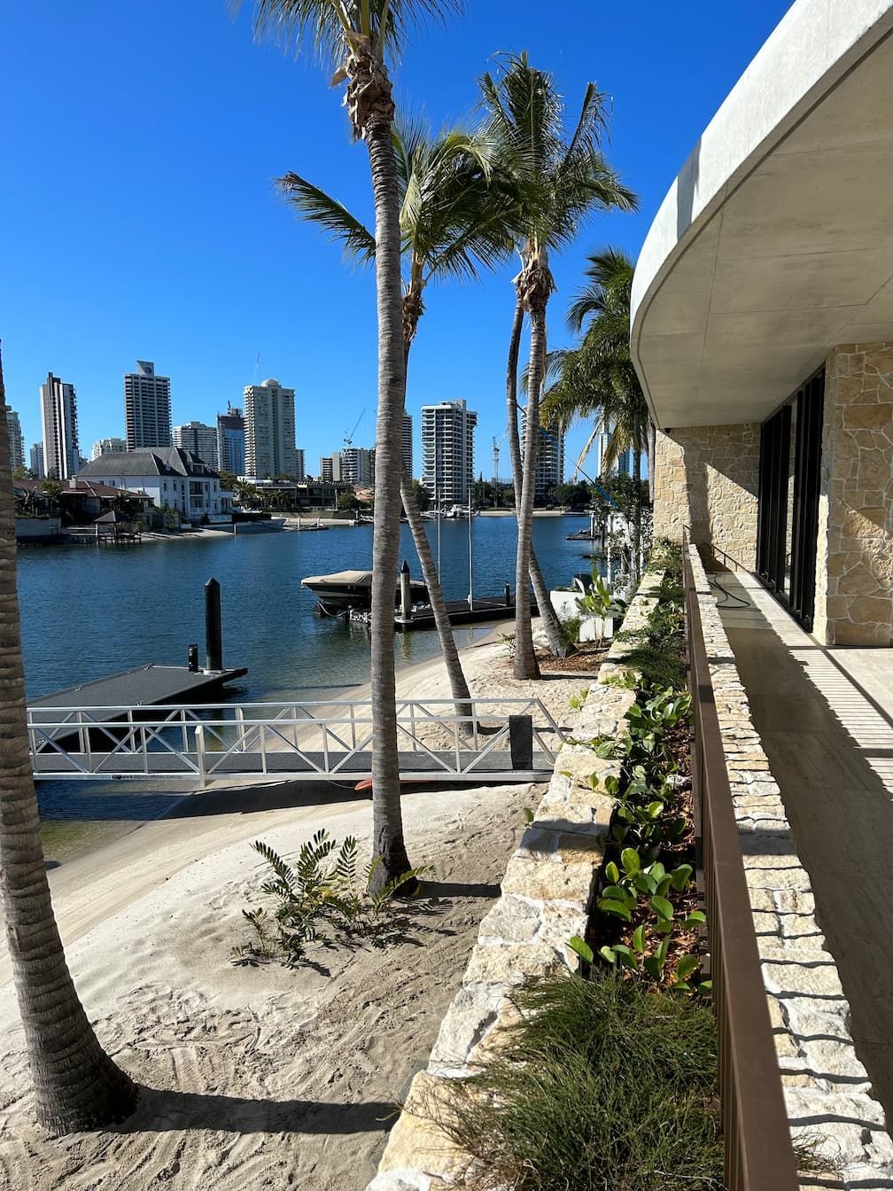 A waterfront view showing a sandy beach, palm trees, a small dock, and a distant city skyline under a clear blue sky.
