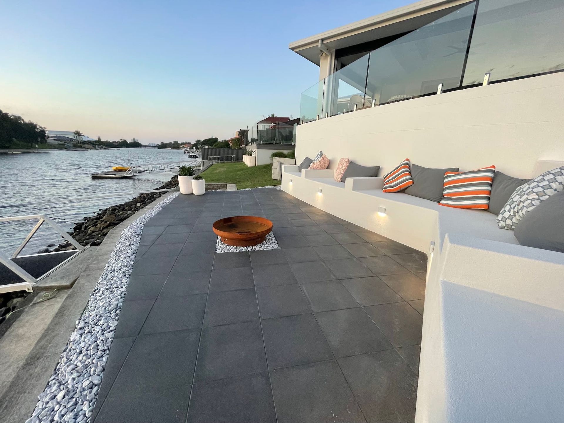 A modern outdoor waterfront patio with dark tiles, a stone fire pit, built-in white seating with cushions, and a glass rail.