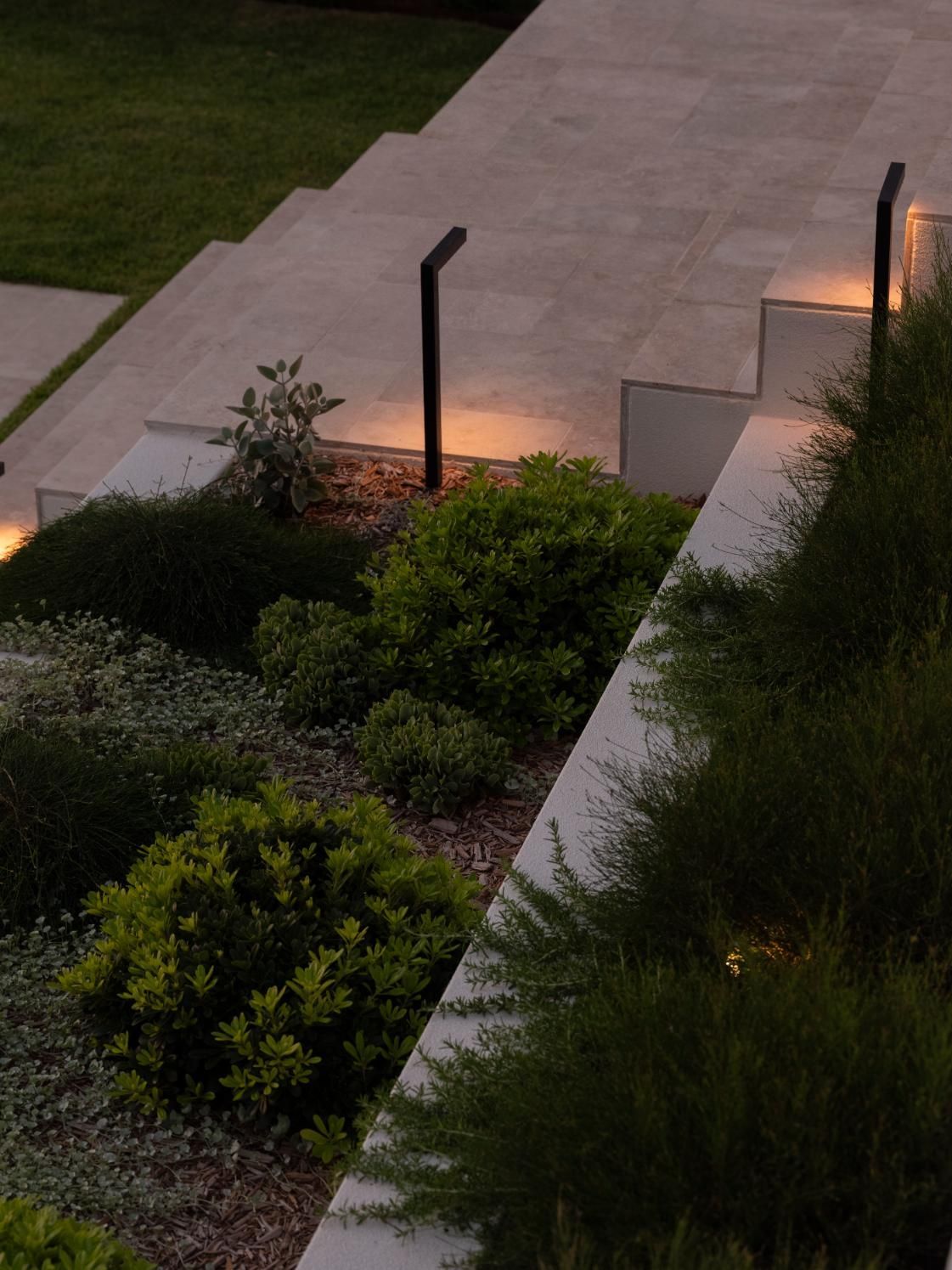 Modern tiered stone steps in a garden at dusk, illuminated by minimalist black pathway lights nestled among greenery.
