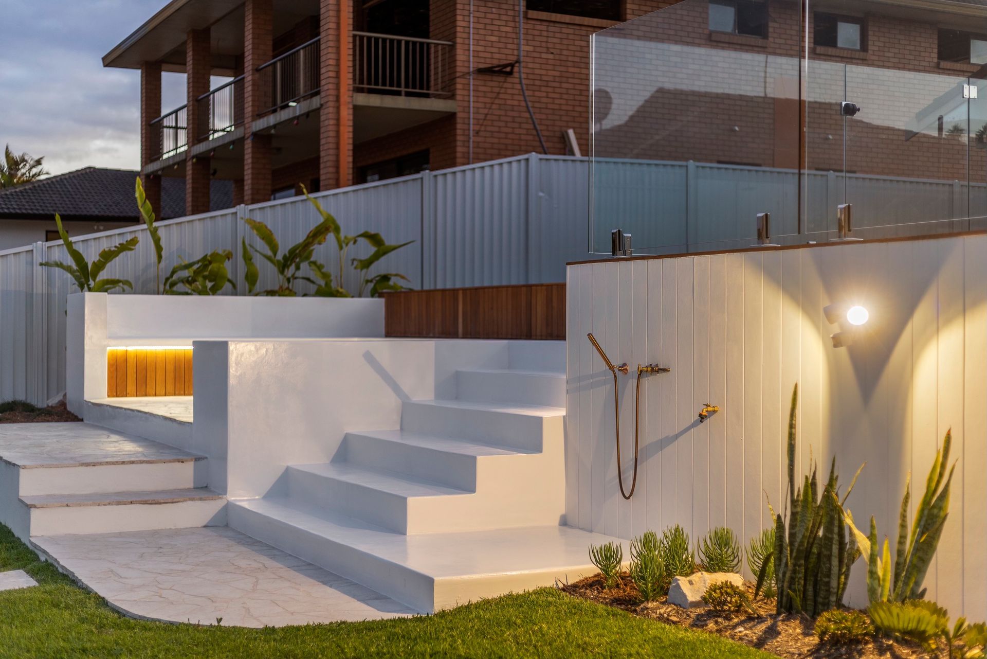 Modern white tiered concrete steps illuminated by warm LED strips, leading to a backyard garden area at twilight.