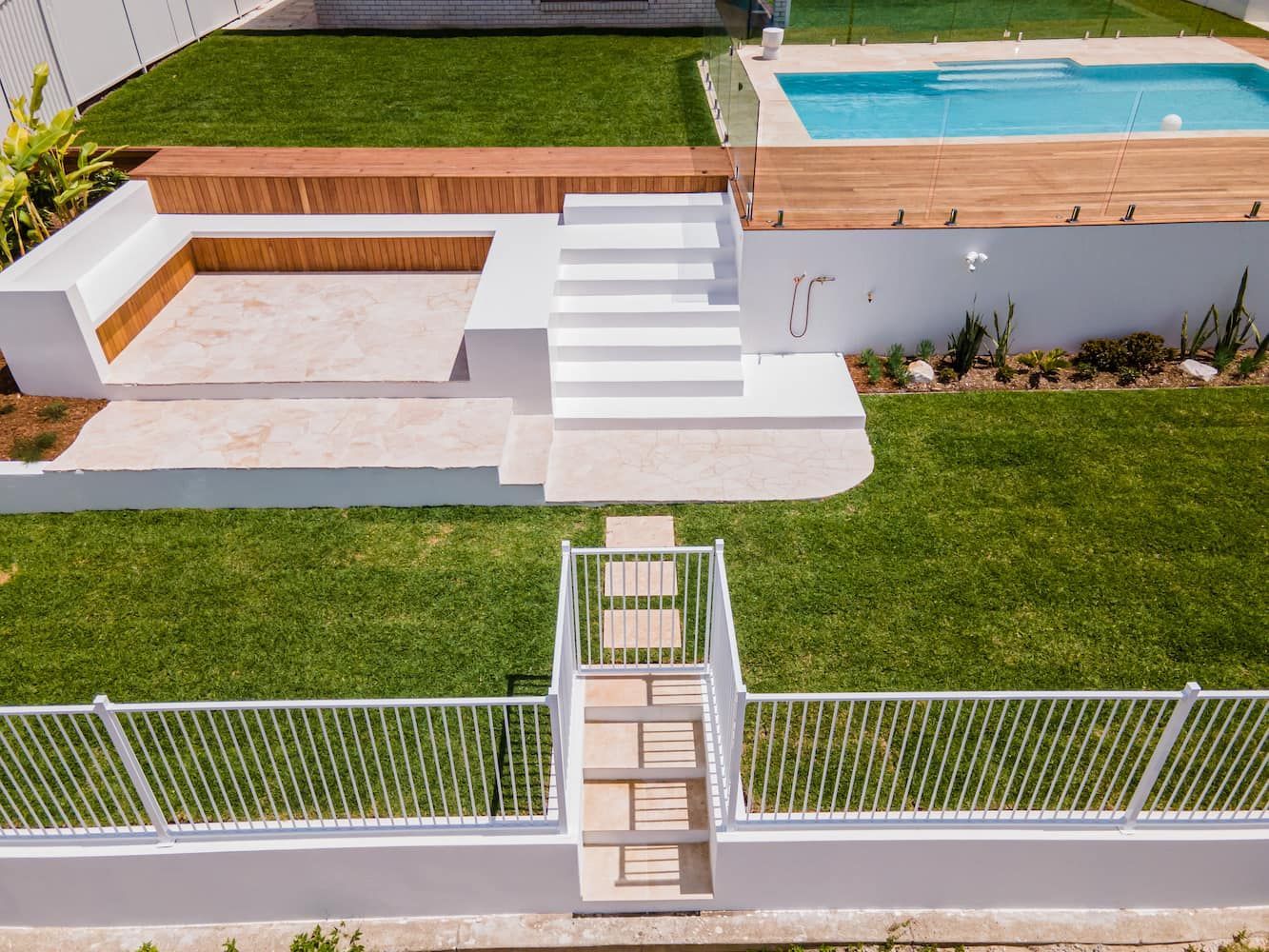 An elevated backyard with a swimming pool, wooden deck, sunken seating area, white stairs, and green lawns behind a fence.