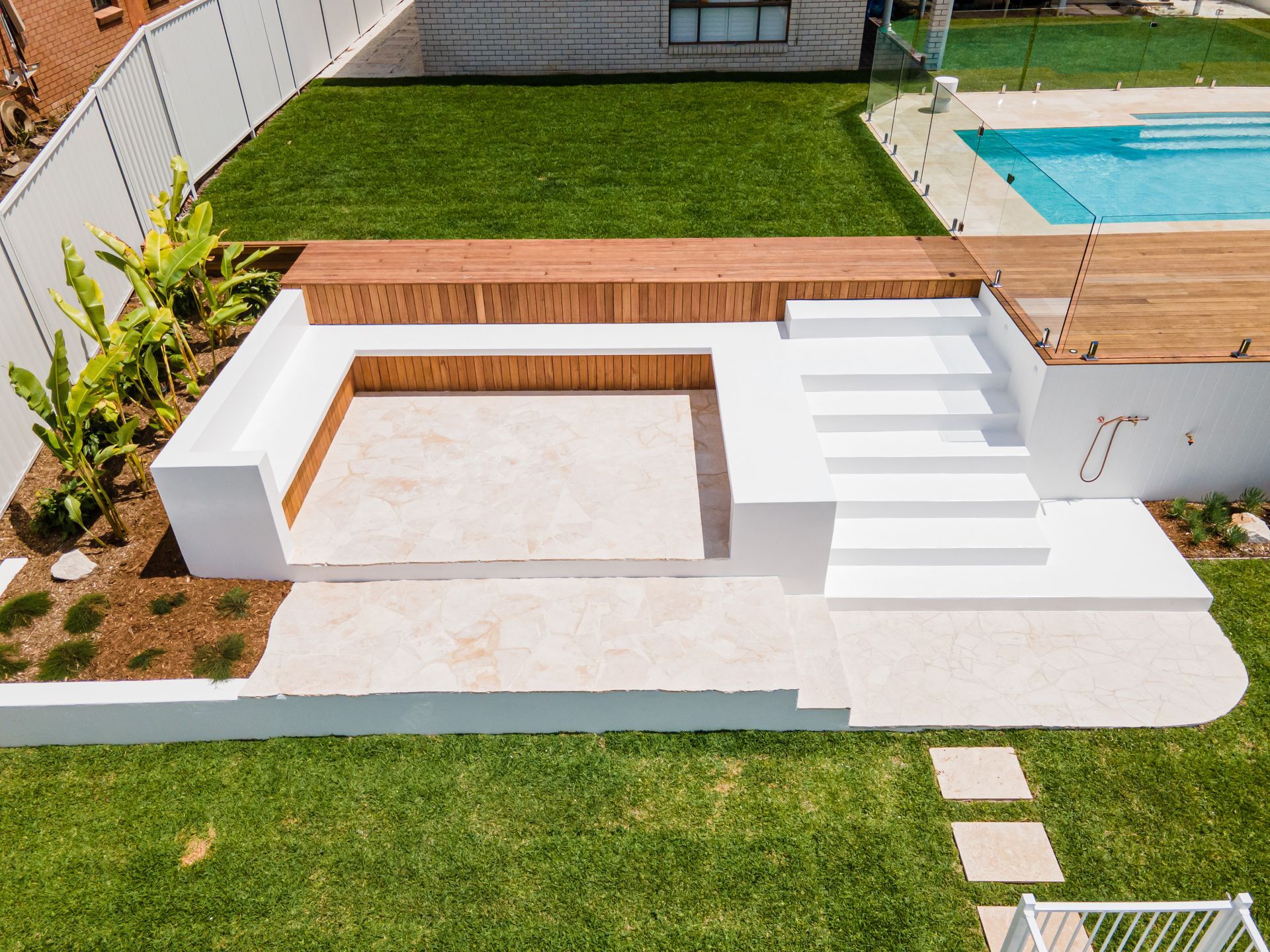 An outdoor sunken seating area with white concrete walls, steps, and light stone flooring next to a wooden pool deck.