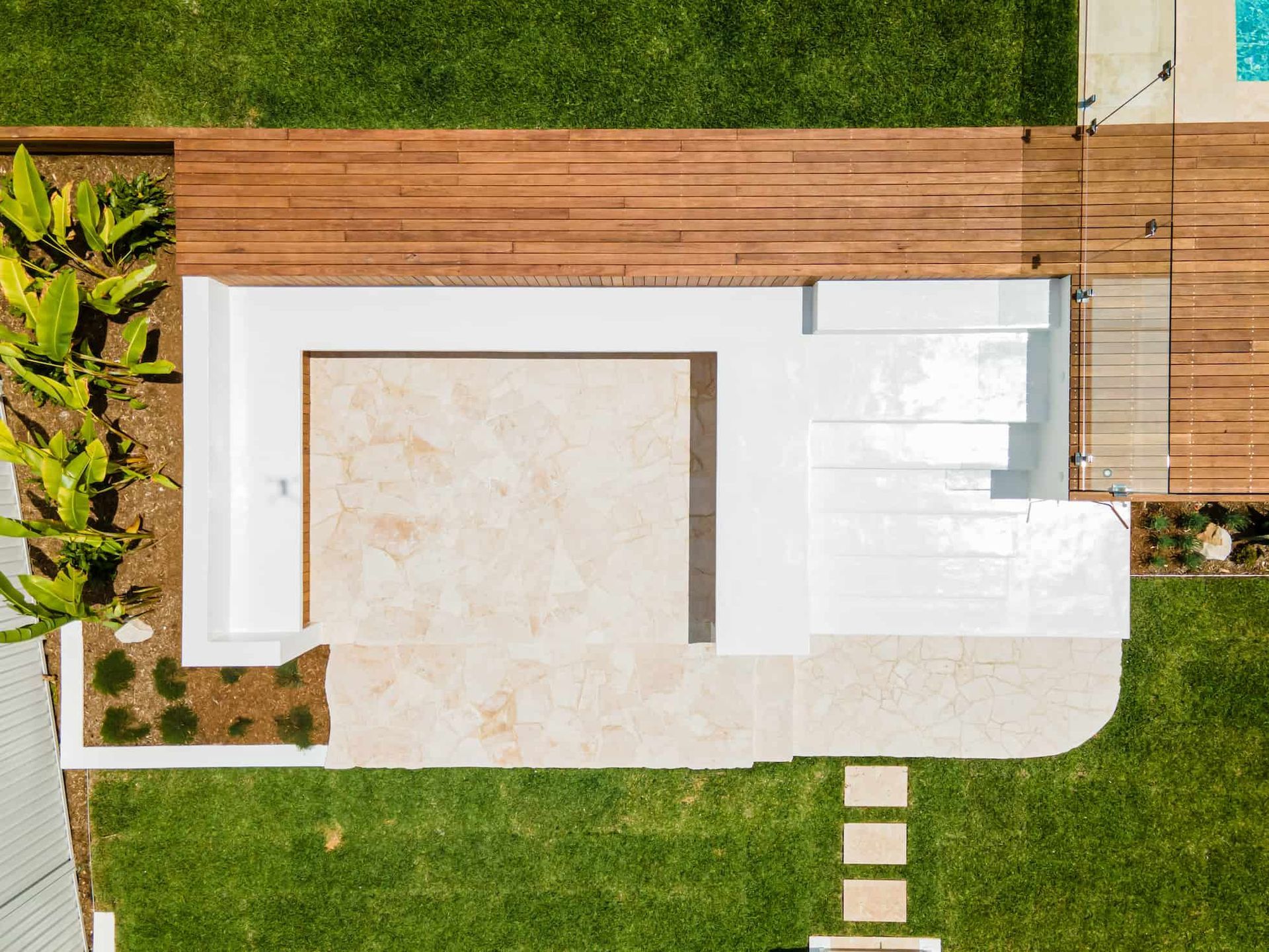 An aerial view of a modern backyard featuring a stone patio, white retaining walls, wooden decking, and green grass.