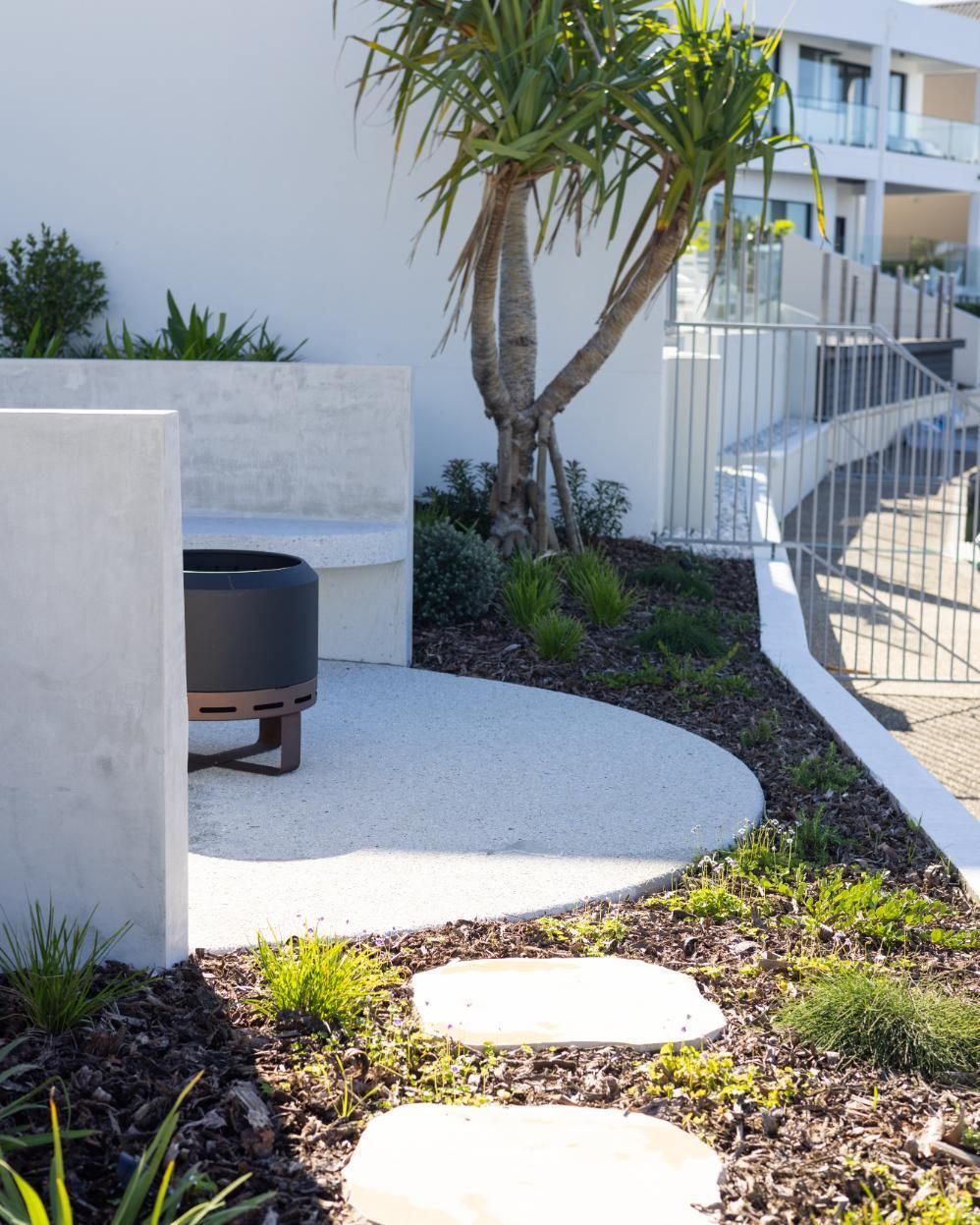A circular patio with a fire pit and concrete seating nestled in a modern landscape with stepping stones and a tree.