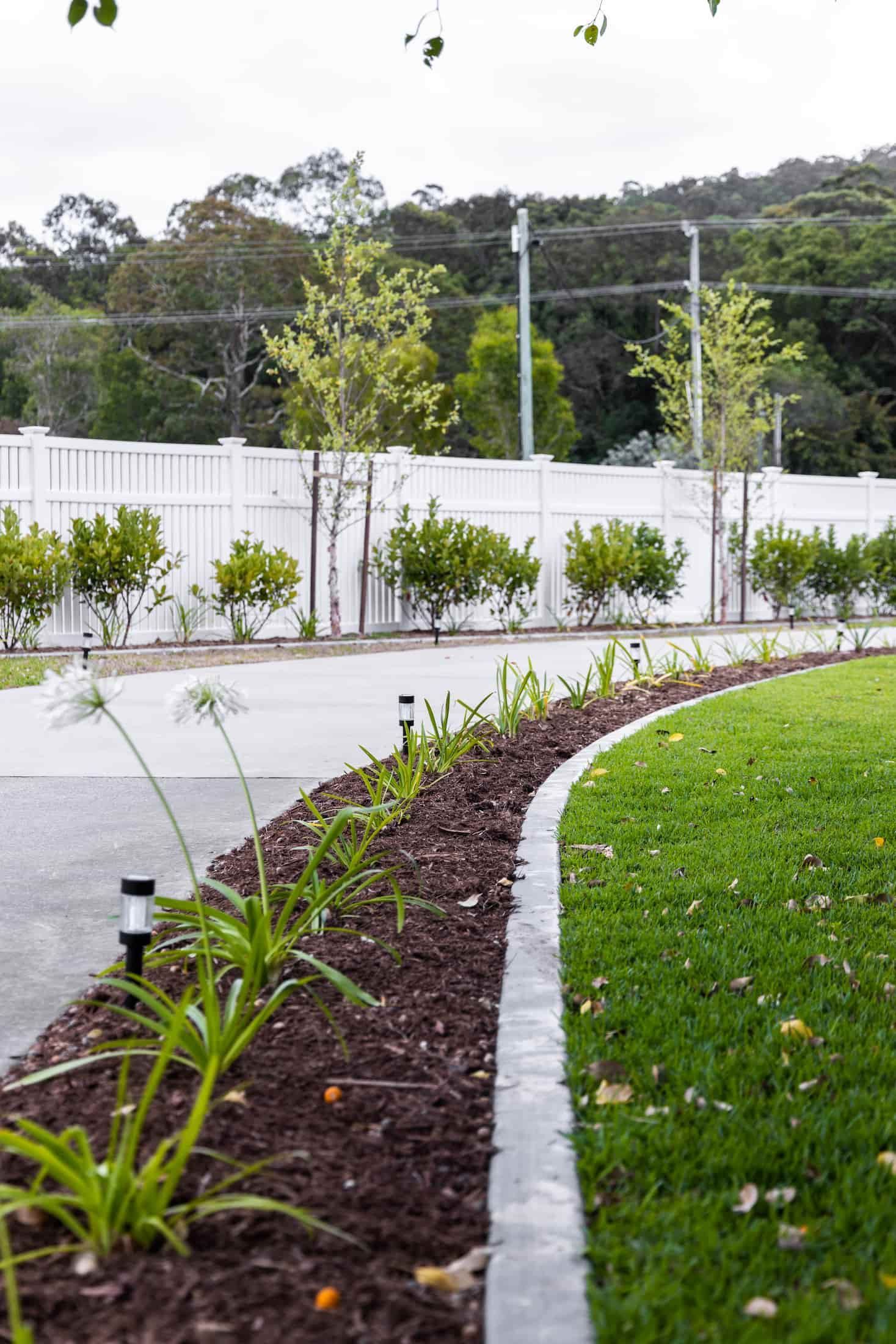 A garden bed with dark mulch and plants, bordered by a concrete edge, next to a curved lawn and a white fence.