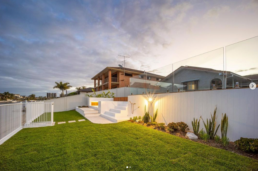 A landscaped backyard at twilight features a bright green lawn, white retaining walls, steps, and a glass-paneled fence.
