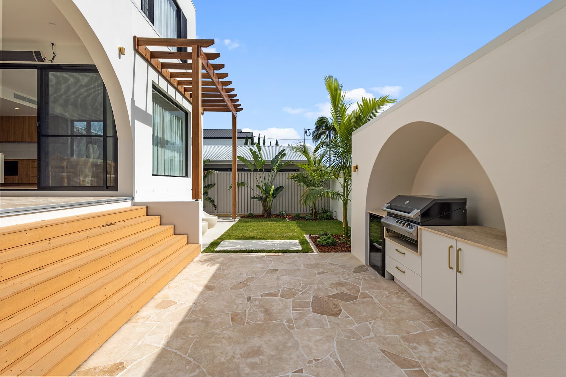A patio with a wooden deck, stone flooring, and an outdoor kitchen built into an arched, white stucco wall.