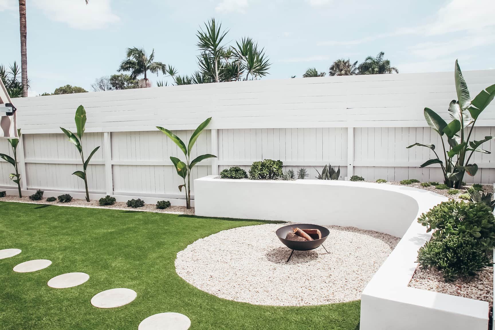 A backyard featuring a white curved wall, a gravel fire pit area, a green lawn with stepping stones, and a white fence.