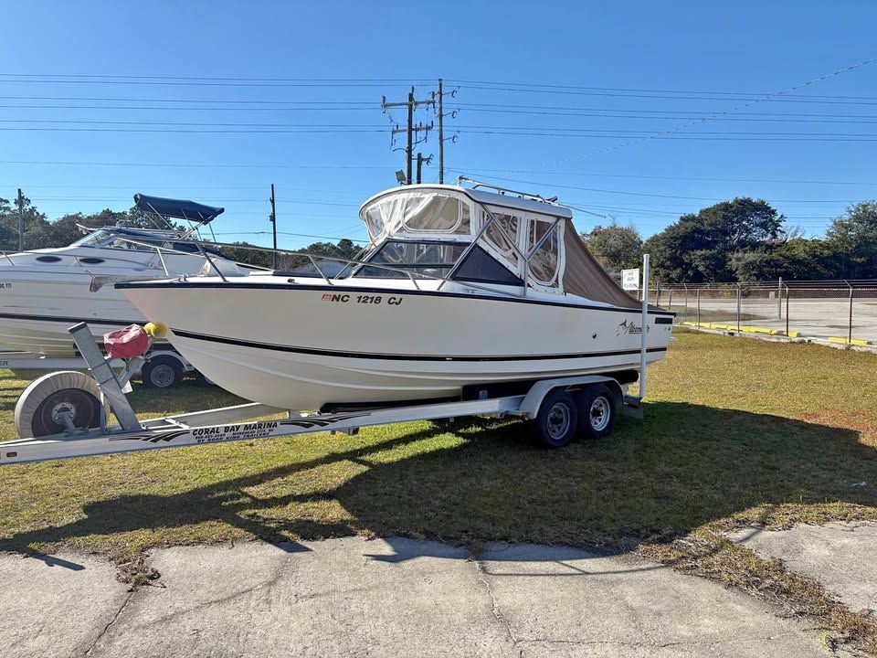 White cabin cruiser boat on a trailer parked on grass near a road under a clear blue sky.