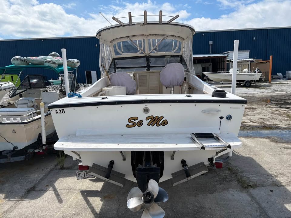 White boat named So Me on a trailer at a boatyard, rear view with beige canopy and twin outboards