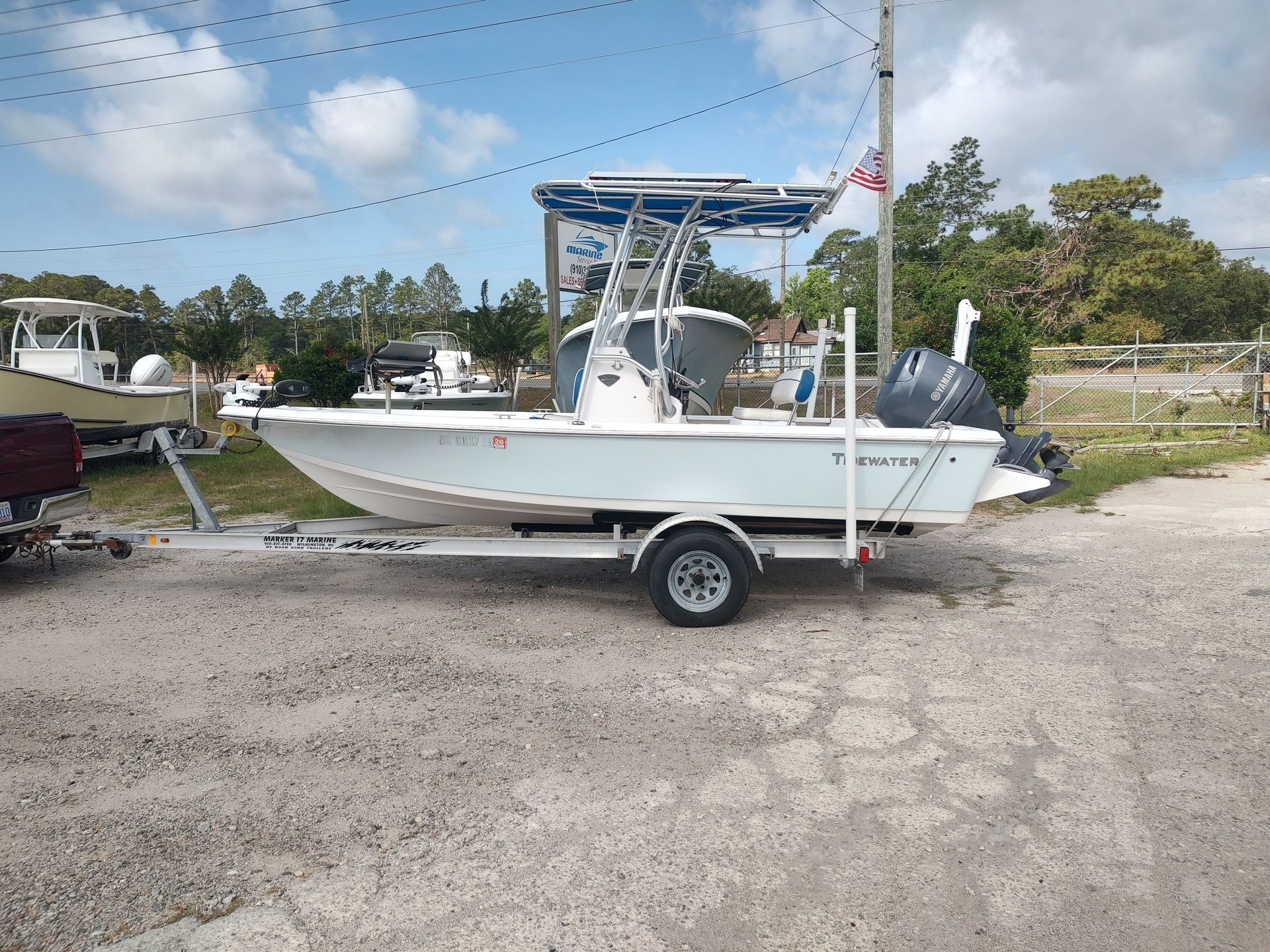 White center-console boat on trailer in a gravel lot under a blue canopy, with an outboard motor.