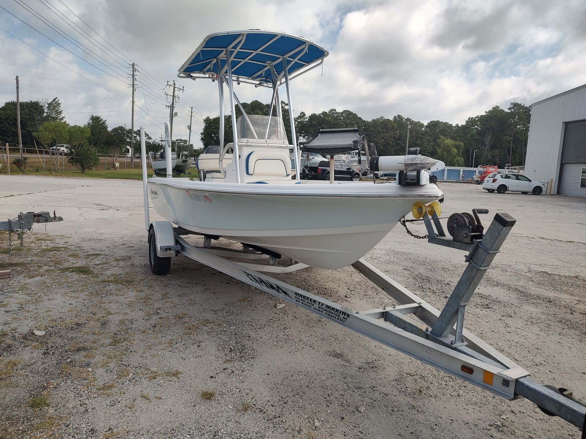 White center-console boat on a trailer in a gravel lot, with a blue canopy and overcast sky