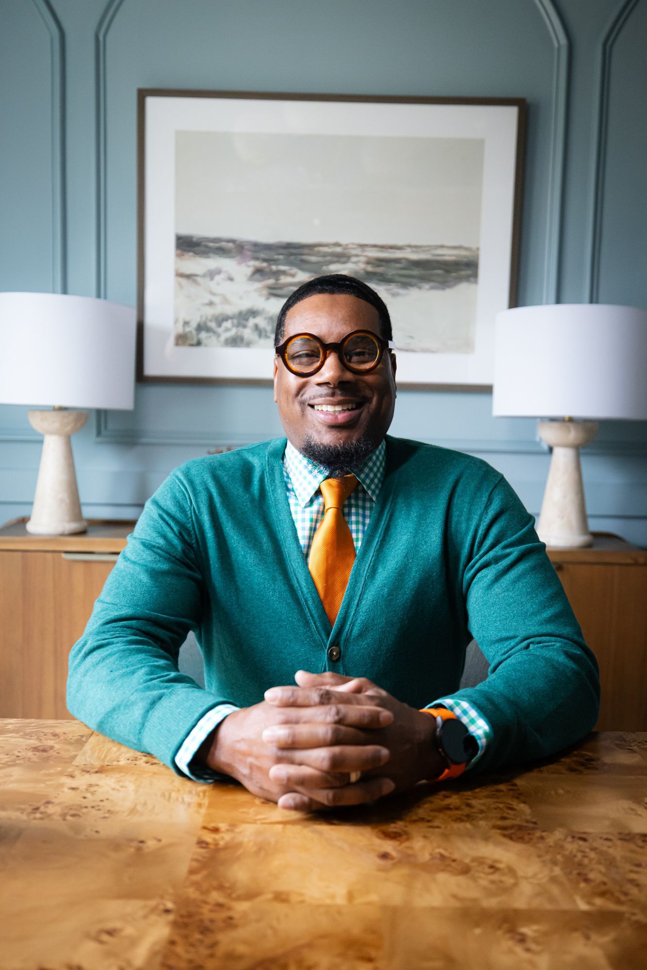 Man smiling at a wooden table, wearing a teal sweater and yellow tie, in a blue room with lamps and framed art