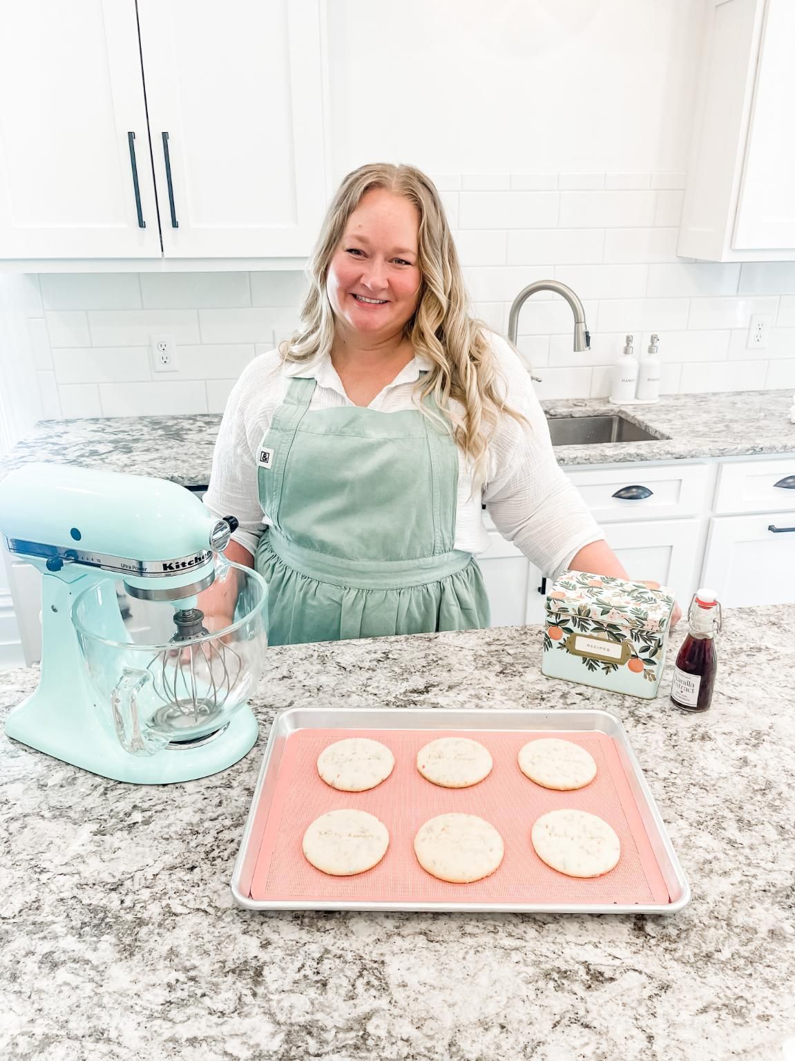 A person wearing an apron stands in a white kitchen behind a counter with a stand mixer and a tray of six cookies.