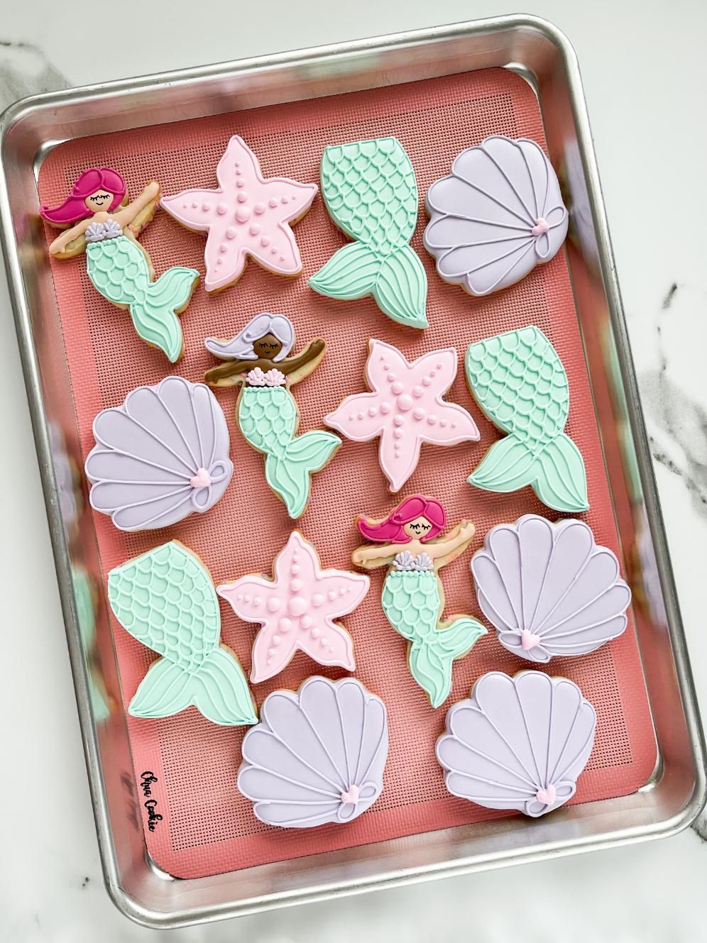A metal baking sheet with mermaid-themed cookies, including mermaids, starfish, and seashells in pastel pink and teal.