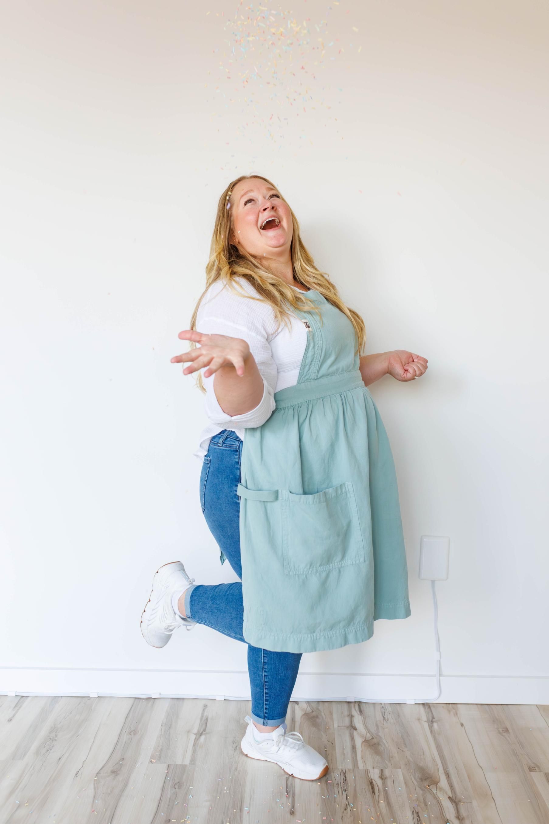 A person wearing a white shirt, blue jeans, and a light blue apron, laughing with one leg raised as confetti falls.