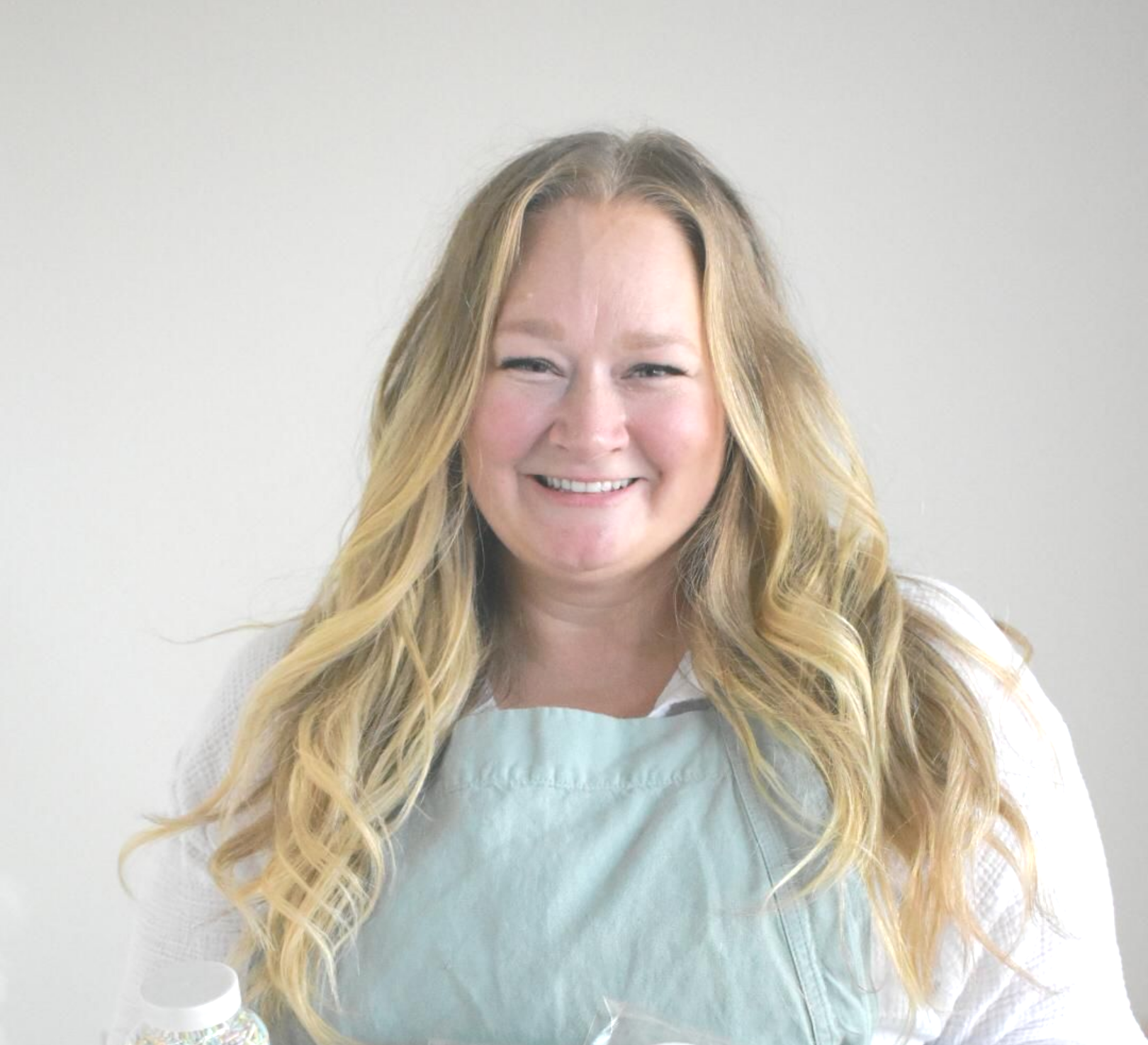 Smiling person with long blonde hair wearing a white shirt and a light green apron against a plain light gray background.
