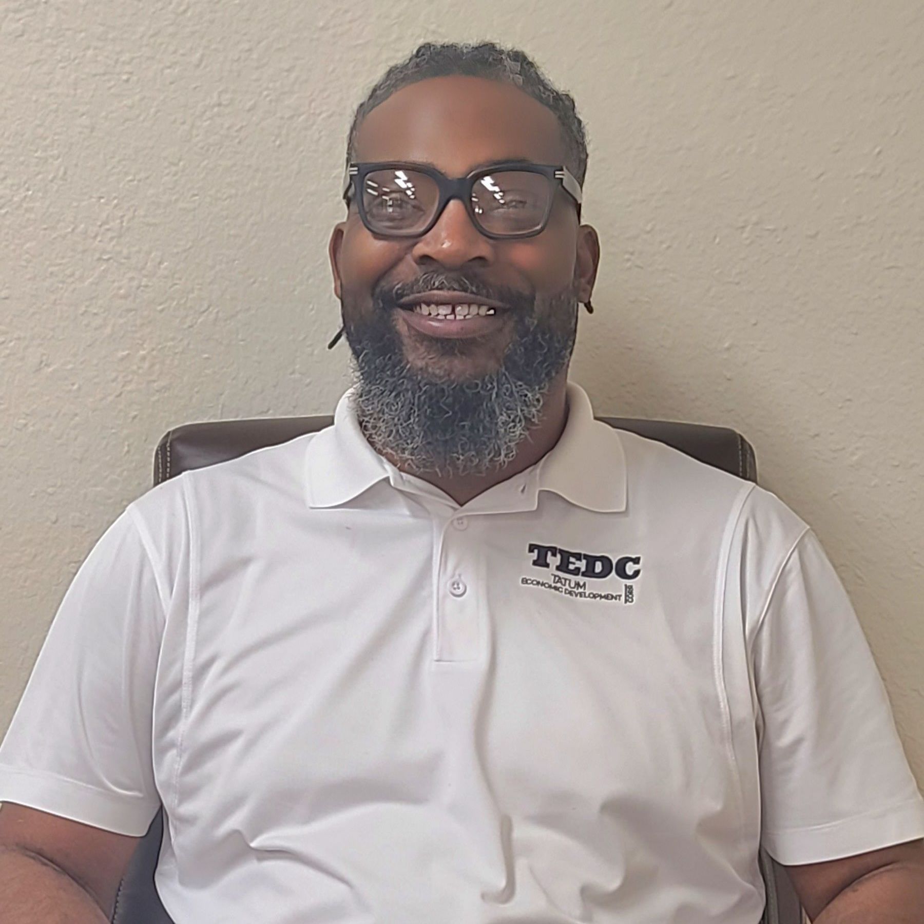 A man with a beard and glasses is wearing a white shirt that says tedc on it.