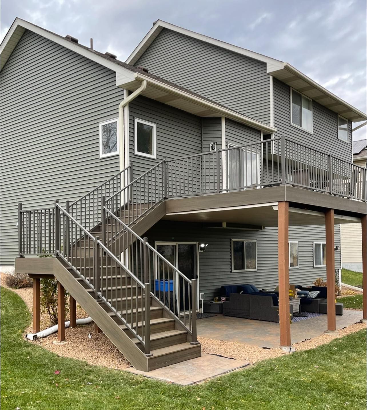 Two-story gray house with deck and stairs. Deck has brown railing. Ground level has patio furniture.