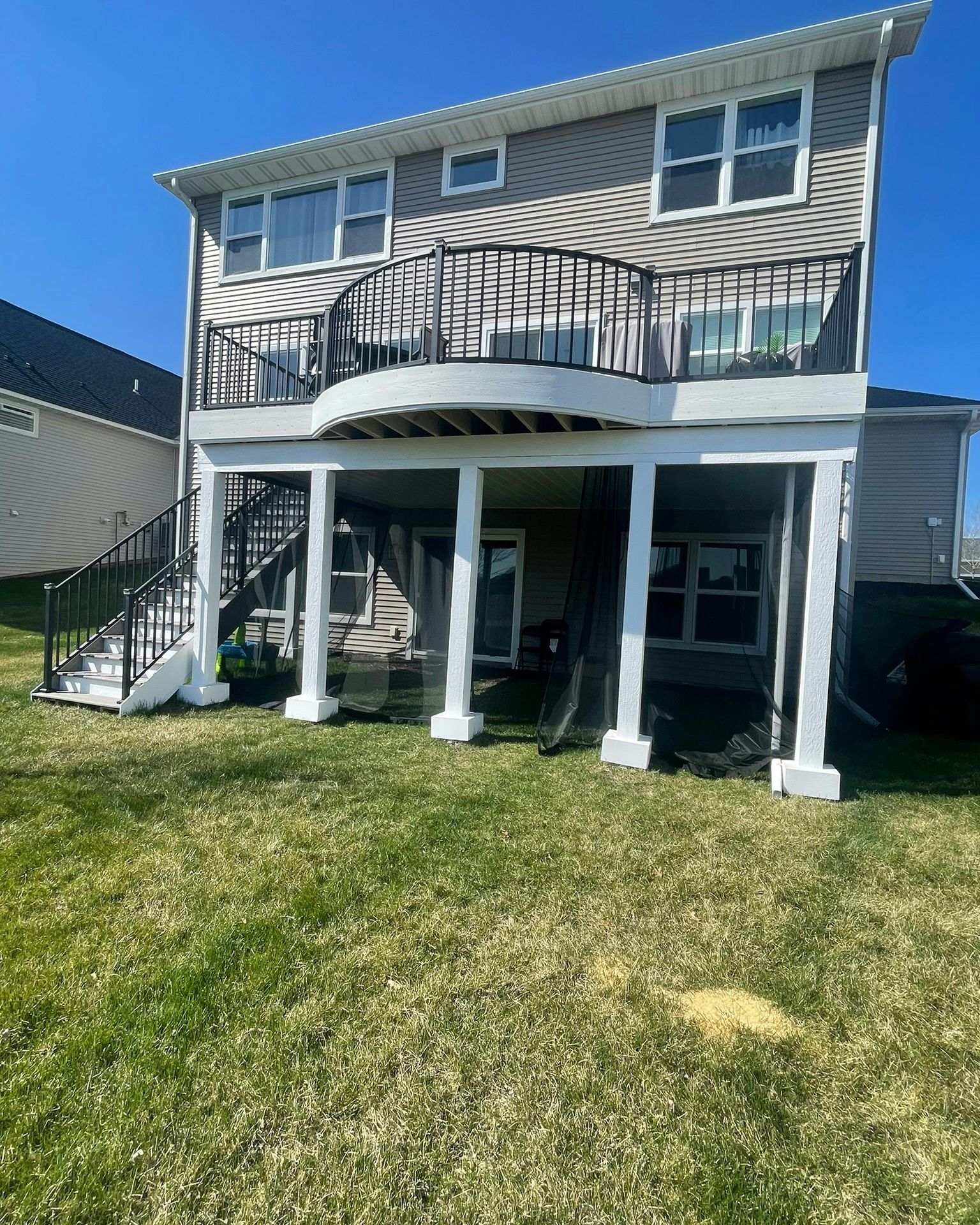 Two-story house with a wooden deck and black railings. White columns support the deck on a grassy lawn under a blue sky.
