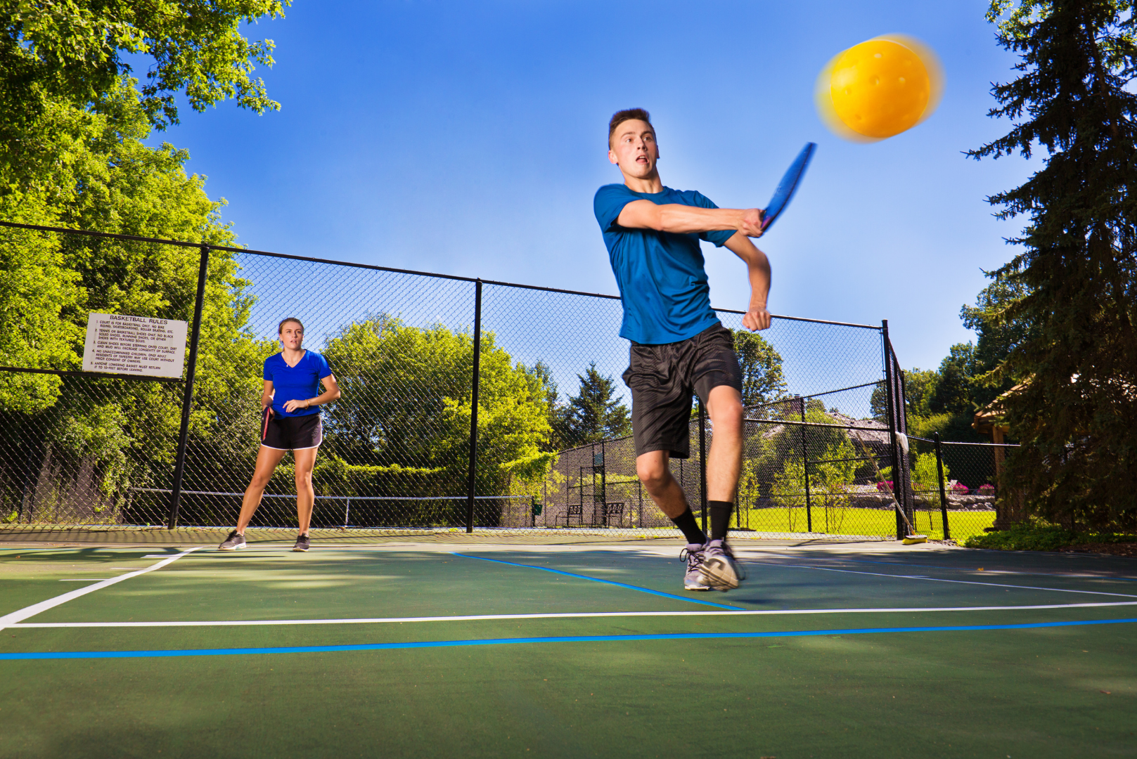 Two players on an outdoor tennis court, one in blue swinging at a yellow ball