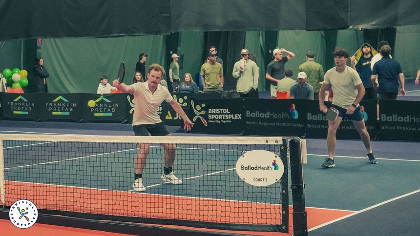 Register Today! Players volley on an indoor pickleball court with spectators behind the net.
