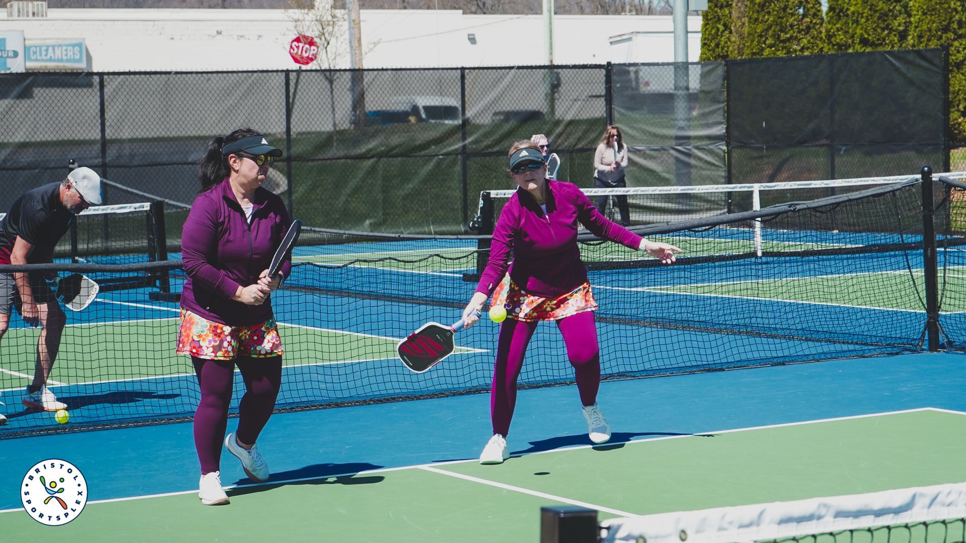 Tennis player in white hitting a backhand at the net, with a yellow ball in motion and sunglasses on.