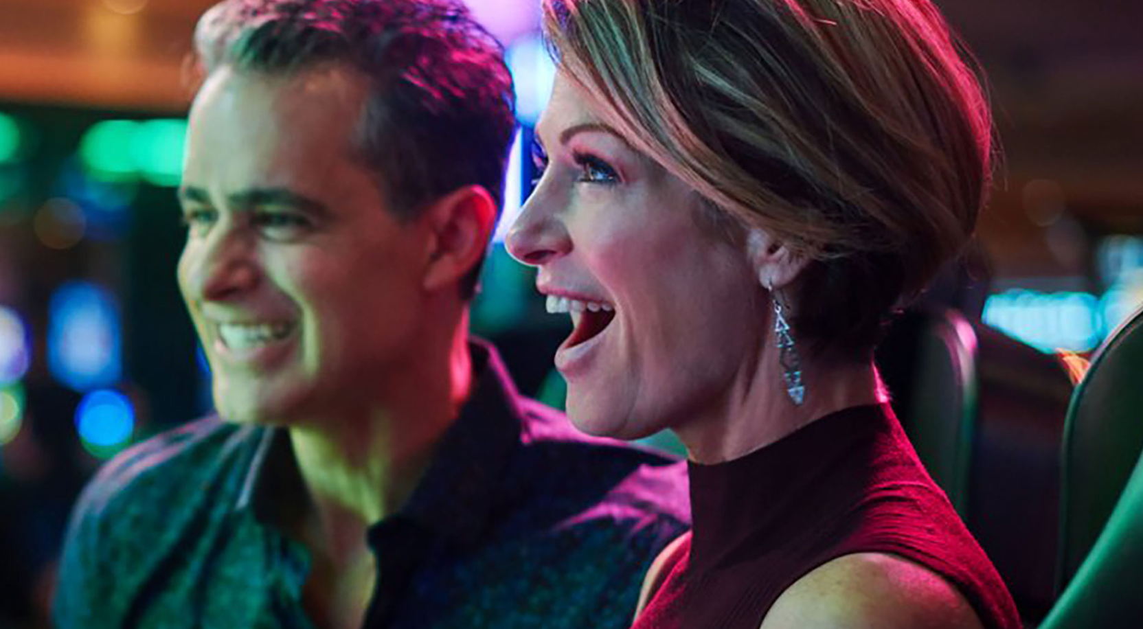 Two smiling people seated in a neon-lit arcade, looking off to the right