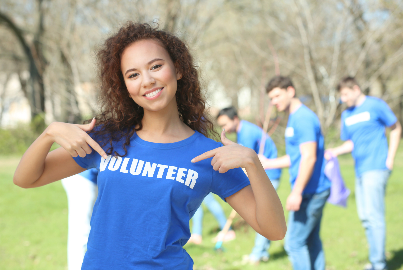 Contact Us to Volunteer Smiling volunteer in a blue shirt pointing at the word “VOLUNTEER” outdoors with others behind her