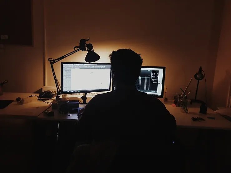 man viewing two computer screens sitting at a desk in the dark