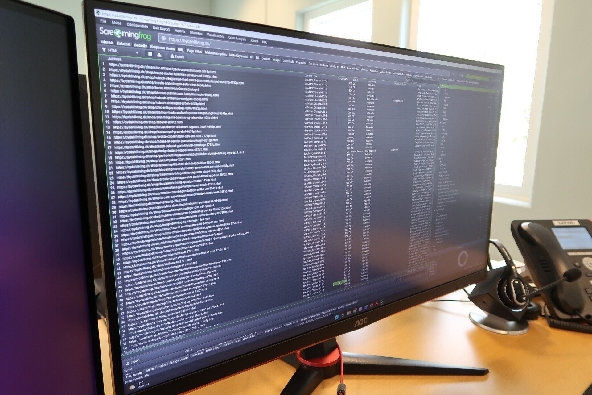 Desktopskærm, der viser et mørkt terminalvindue med tekstkode, ved siden af ​​en bordtelefon på et træbord.