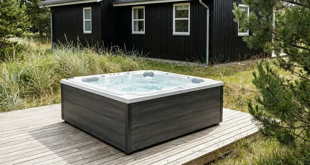 Et firkantet, gråt spabad står på en træterrasse foran et mørkt træhus omgivet af grønne områder.
