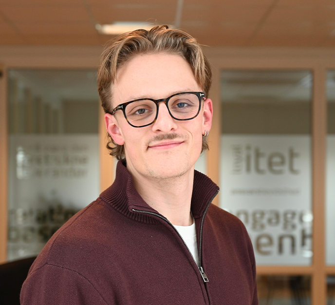 Person med briller og overskæg i en rødbrun sweater, stående indendørs foran glasdøre.