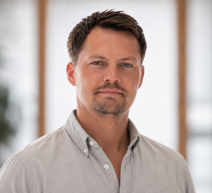 Mand i en lysegrå skjorte med knapper står indendørs og kigger let til siden.
