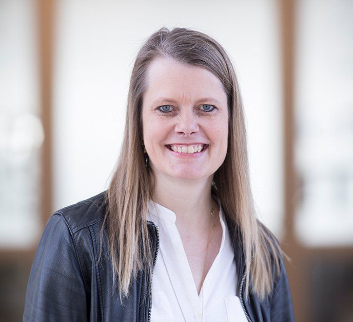 Smilende kvinde i en sort jakke og hvid bluse stående mod en lys, sløret baggrund